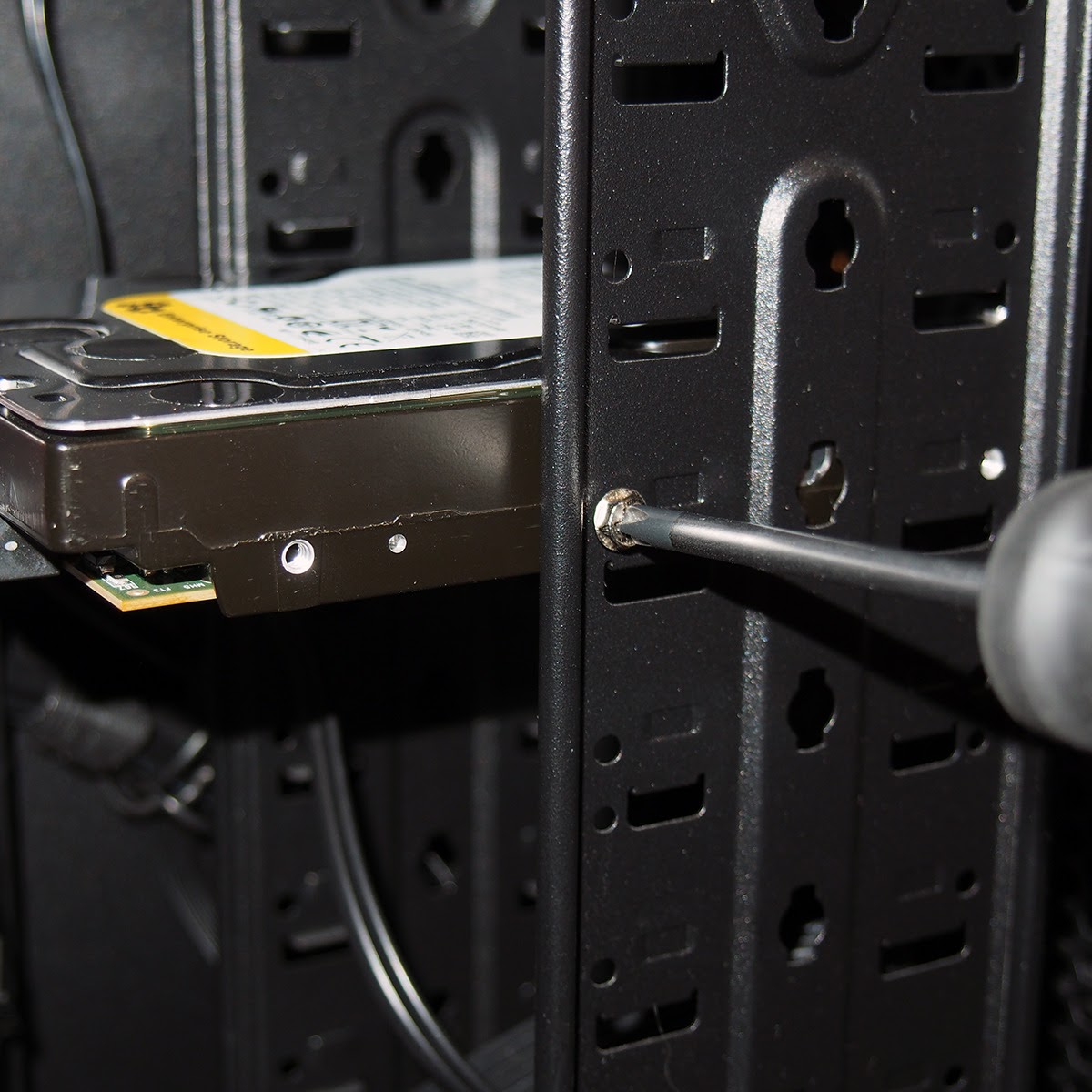 Someone holding a screwdriver and rotating it counterclockwise to replace a Hard Disk Drive inside a computer case. The hard drive is silver, and the case has a matte black finish.