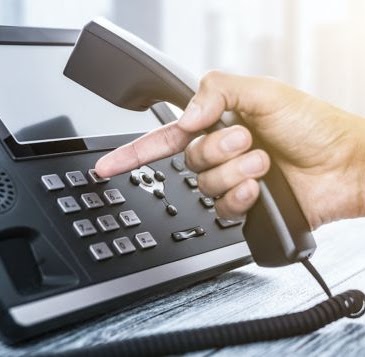 A human hand pictured dialing the number three on the telephone. The phone is placed on a wooden table.